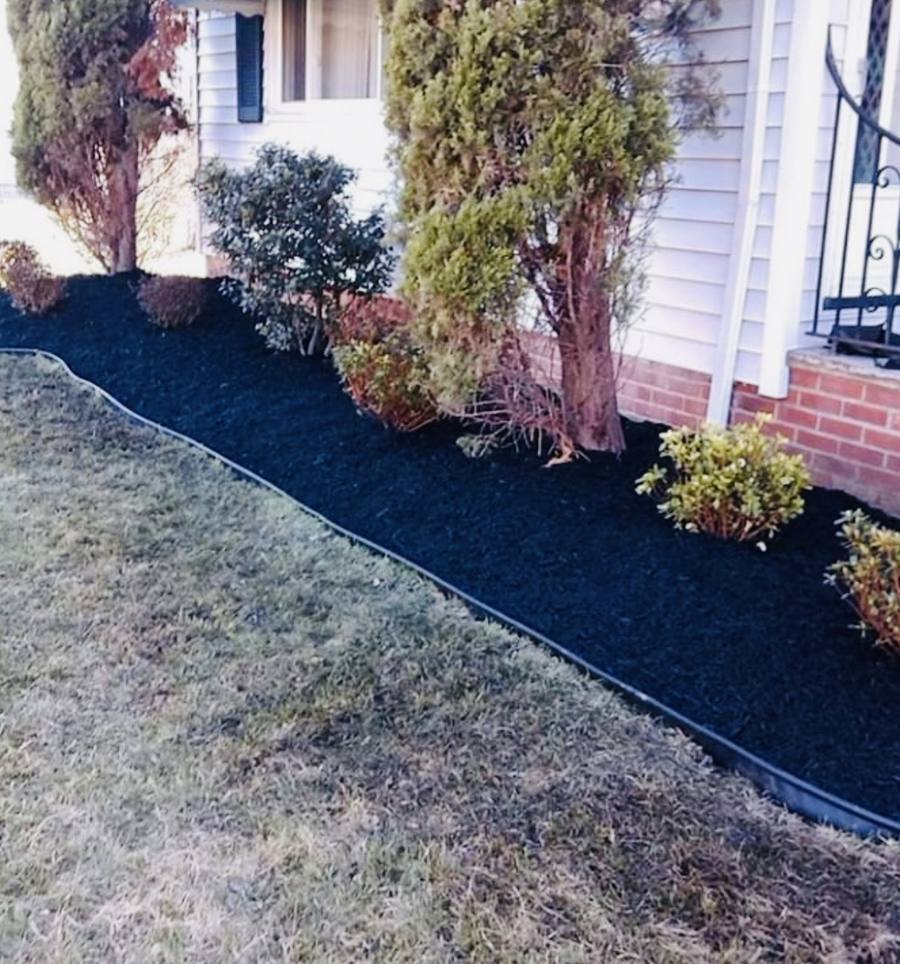 A bed of black mulch edged with a black border strip along the front of a white house, with shrubs behind it and a strip of lawn in front.