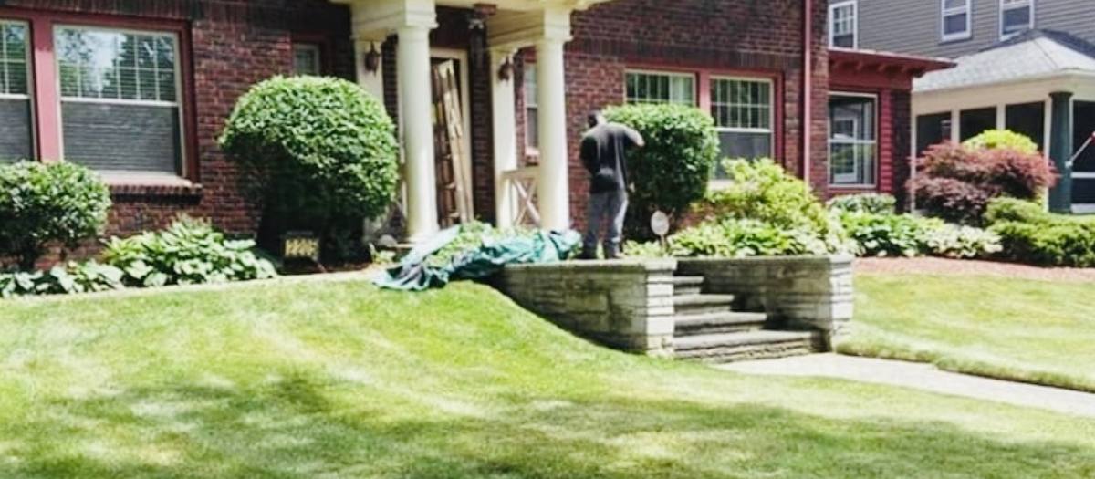 A brick house with white porch columns, clipped round shrubs and hostas along the foundation, a stone-walled front step and a mown lawn; a worker stands in the beds near the steps.